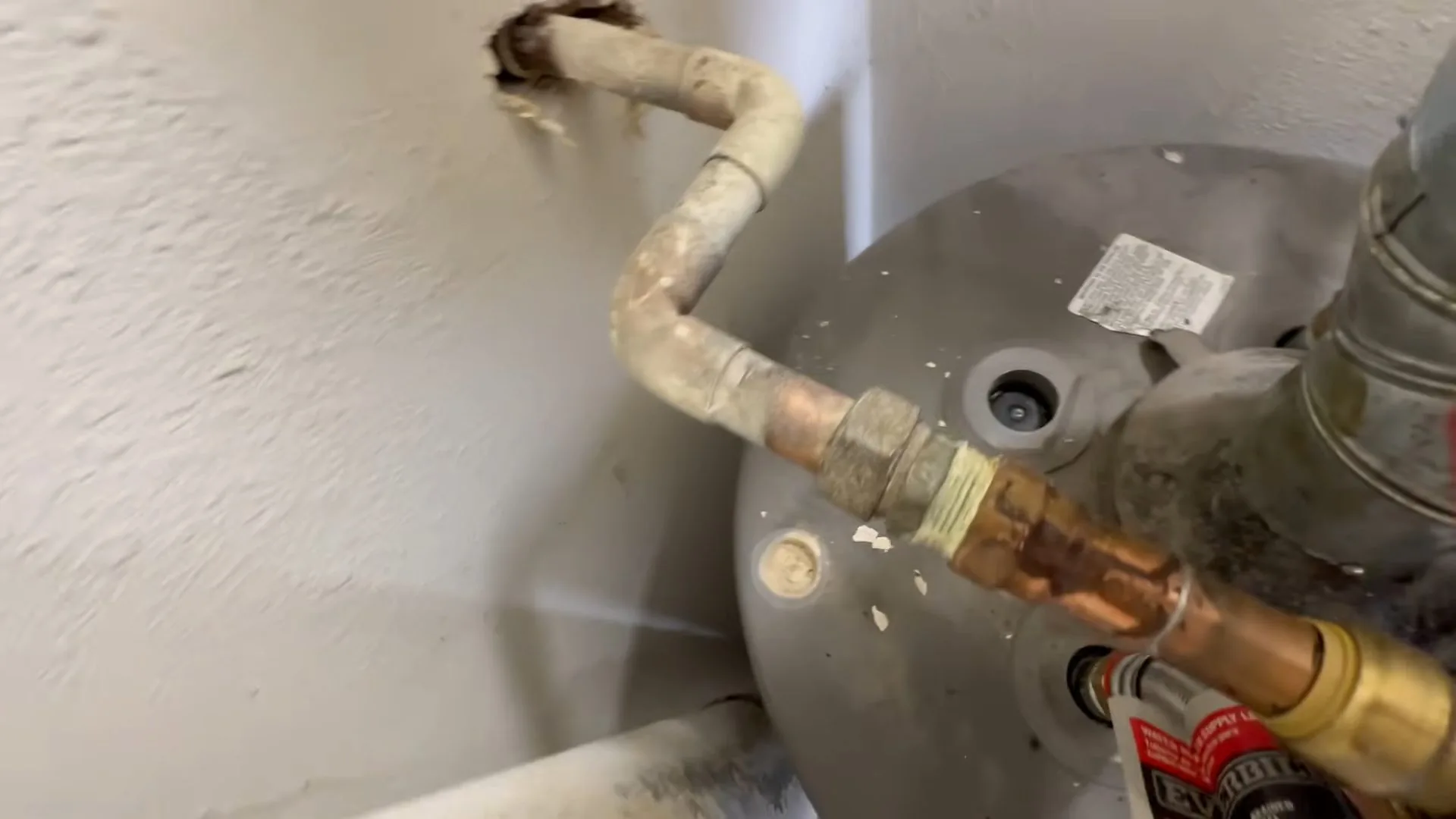 A copper and brass supply line rising from the top of a gray water heater tank beside a hole cut in the wall.