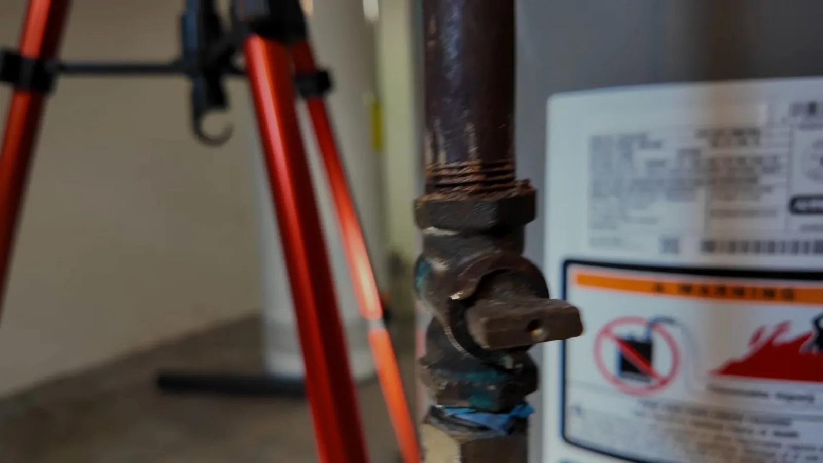 A corroded dark gas shutoff valve beside a water heater label and a camera tripod.