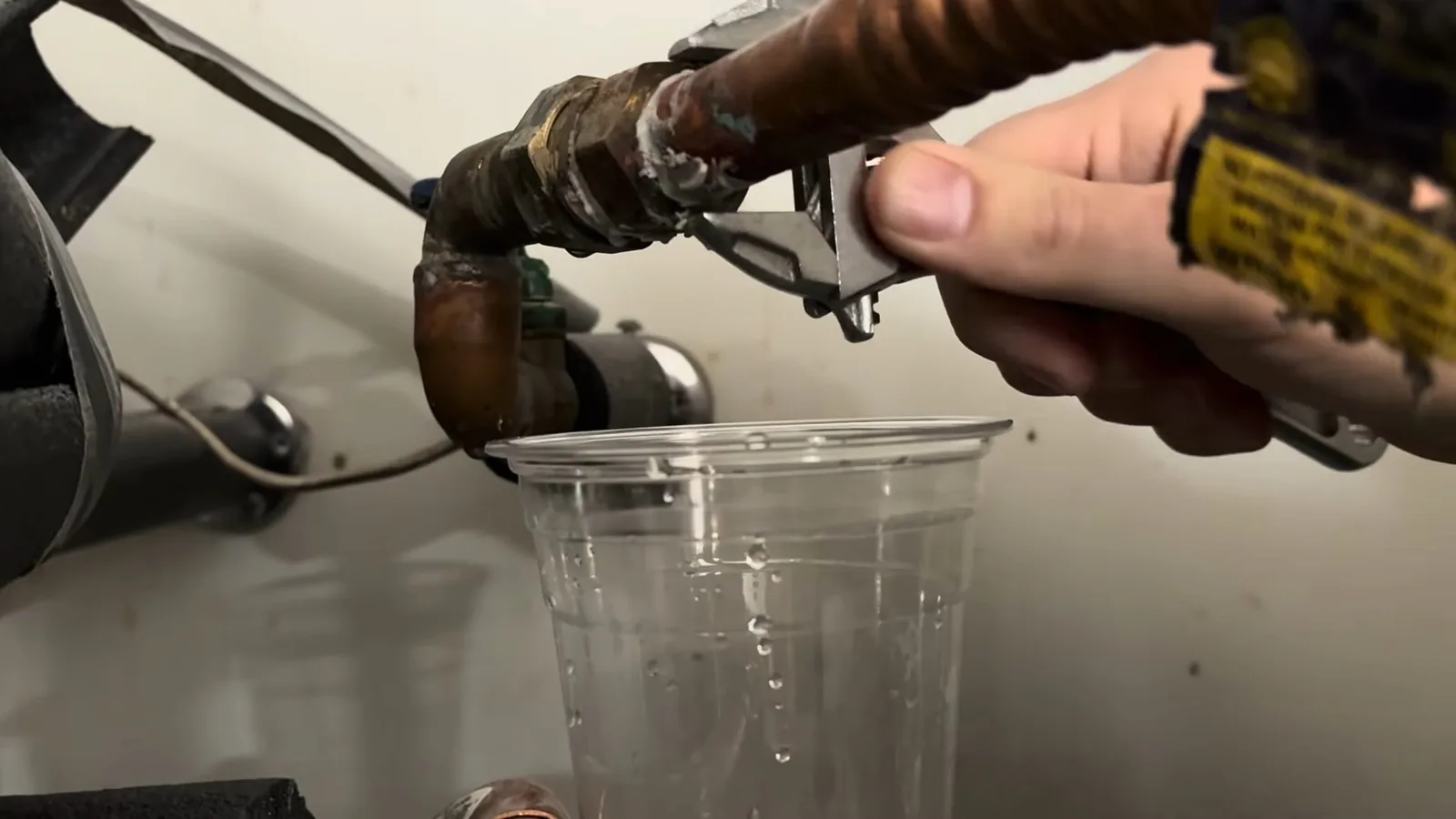 A hand turns an adjustable wrench on a copper fitting at a water heater while water drips into a clear plastic cup.