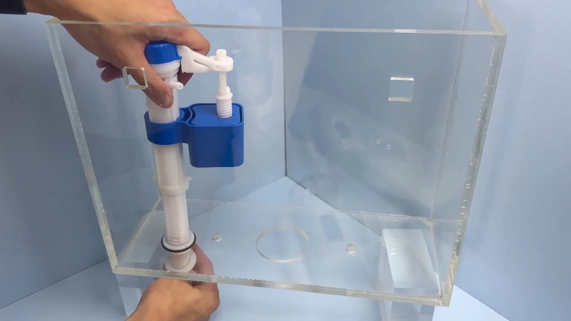 Two hands holding a fill valve and a flush valve inside a clear acrylic toilet tank.