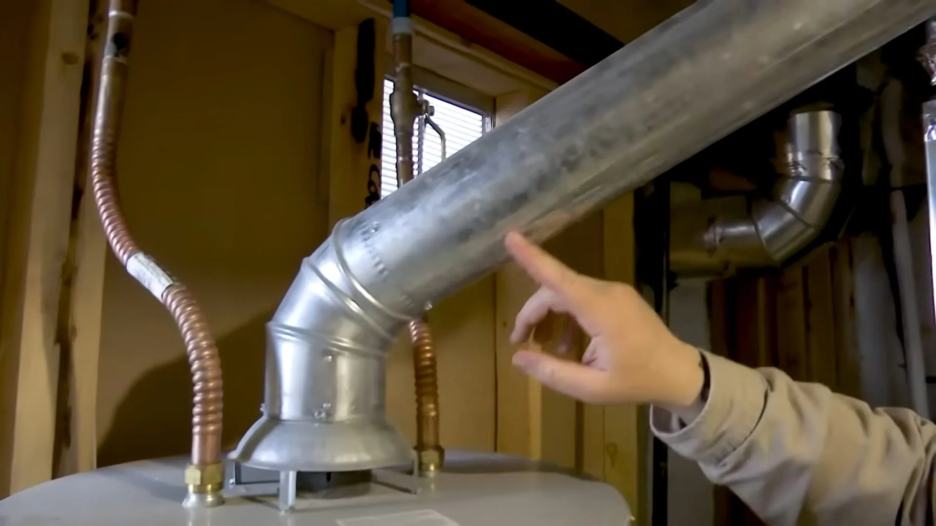 A finger pointing at a galvanized flue elbow on a tank water heater with flexible gas and copper lines.