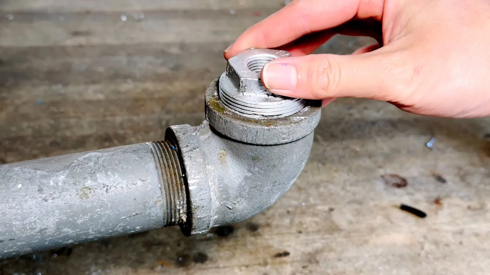 A hand holding a galvanized cleanout plug above a pipe elbow.