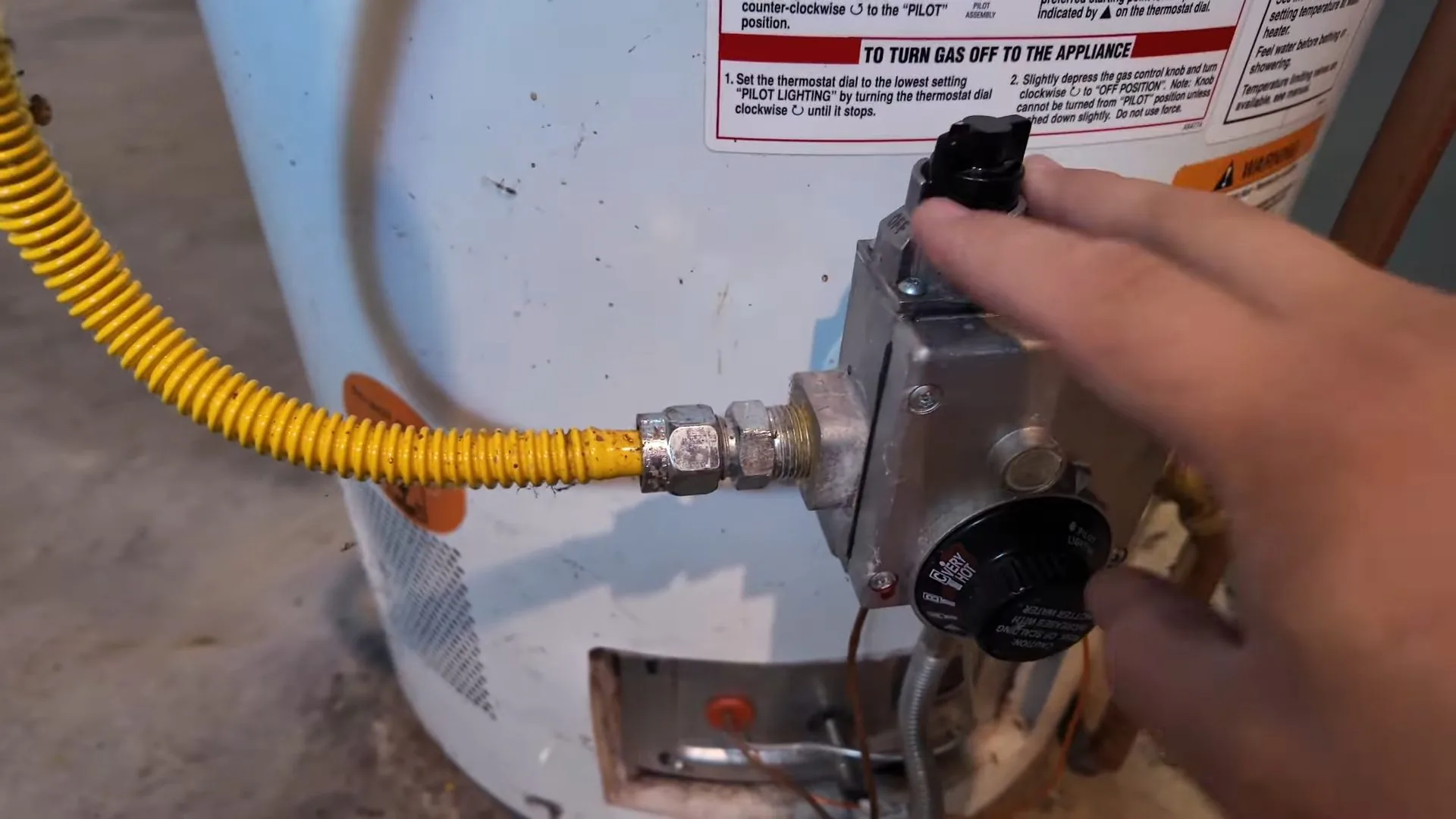 A hand resting on a water heater gas control, with a yellow flexible gas line running to it.