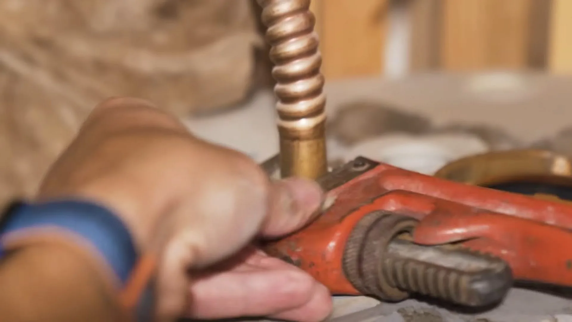 A hand holding an orange pipe wrench on a flexible copper connector at a water heater.