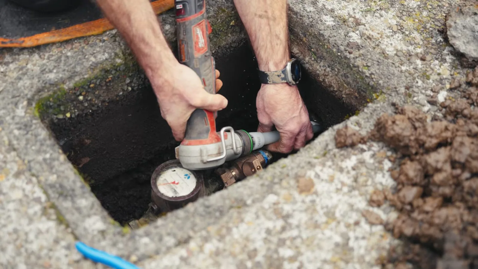 Hands working with a power tool on a service line at a meter inside a ground box.