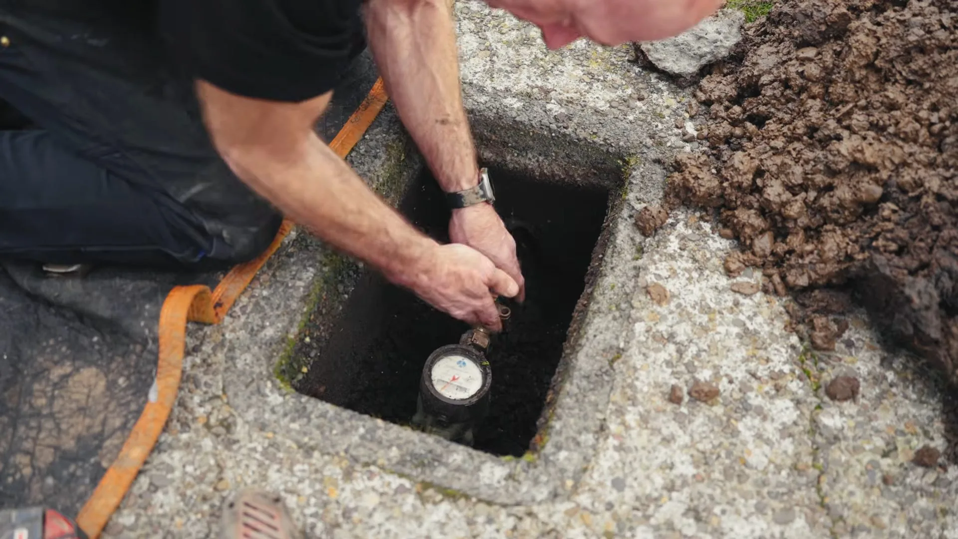 A man kneeling beside a ground meter box and reaching in to read the water meter.