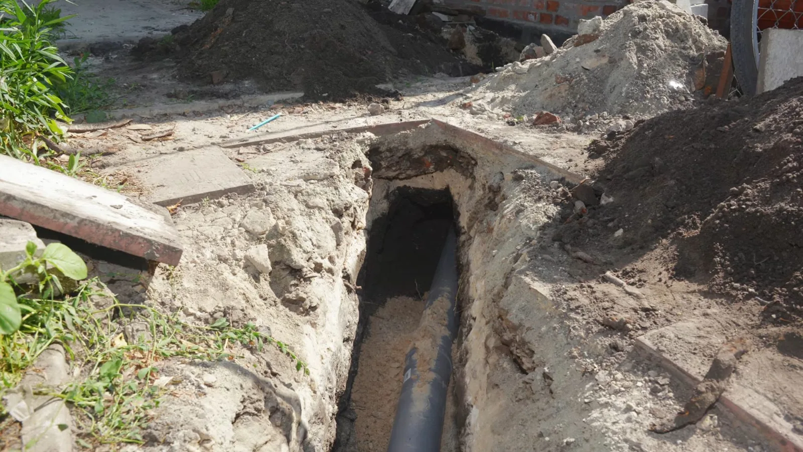 An open trench beside a house exposing a dark sewer pipe.