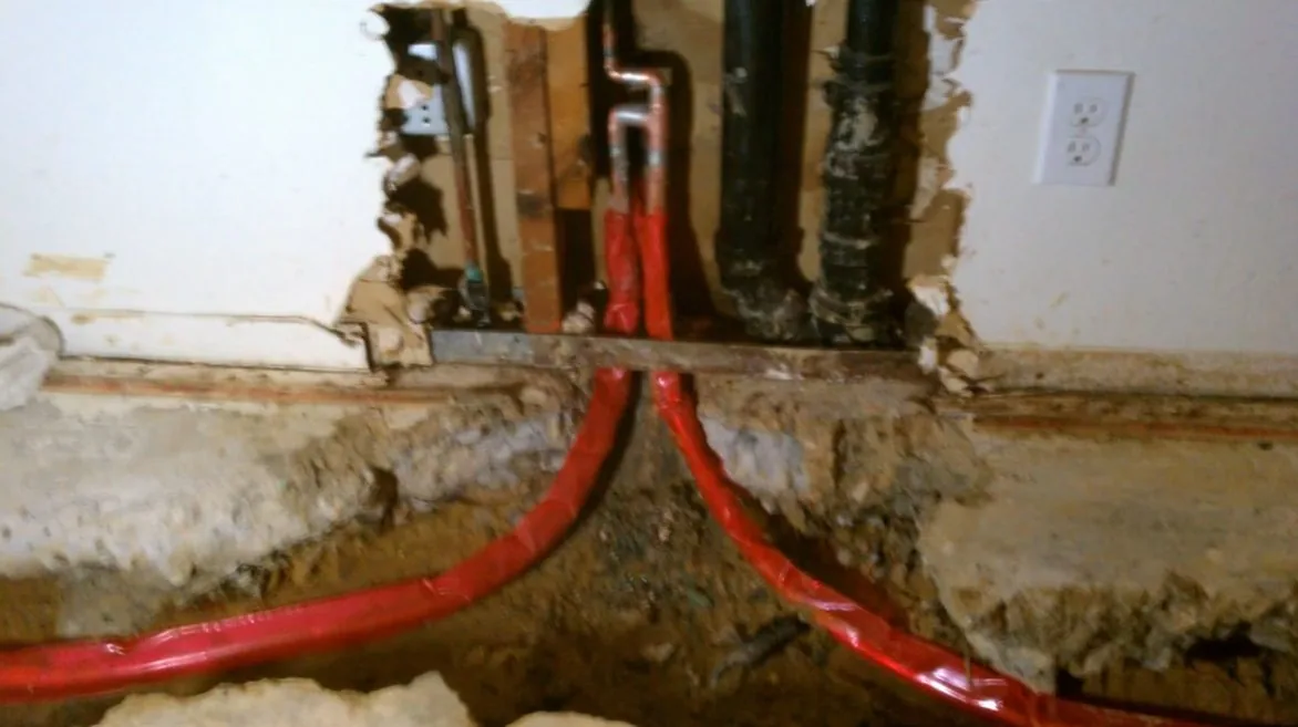 The base of an opened wall showing a rotted bottom plate, red PEX lines and black drain pipes.