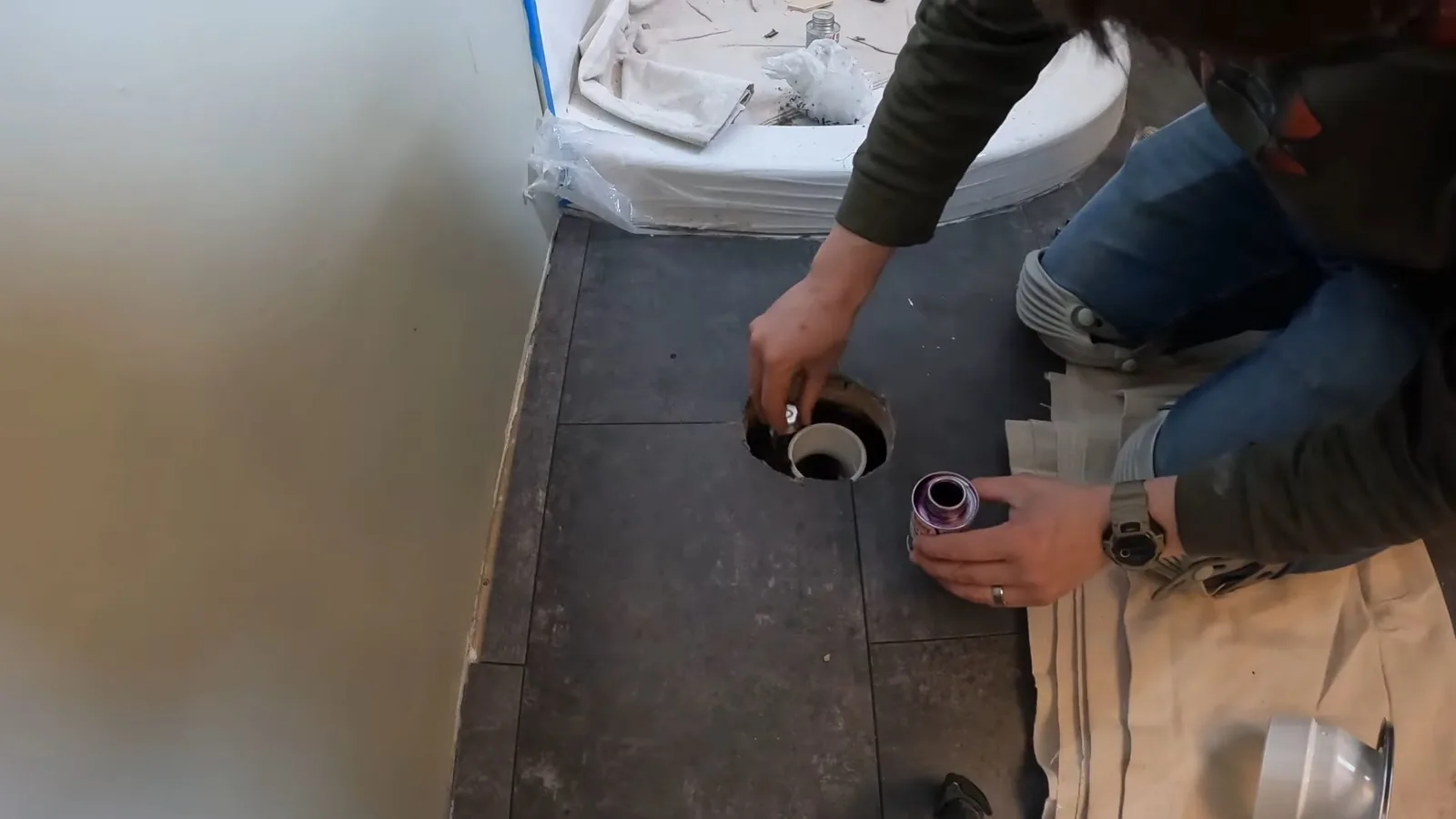 A person kneeling beside an exposed toilet flange on a dark tile bathroom floor.