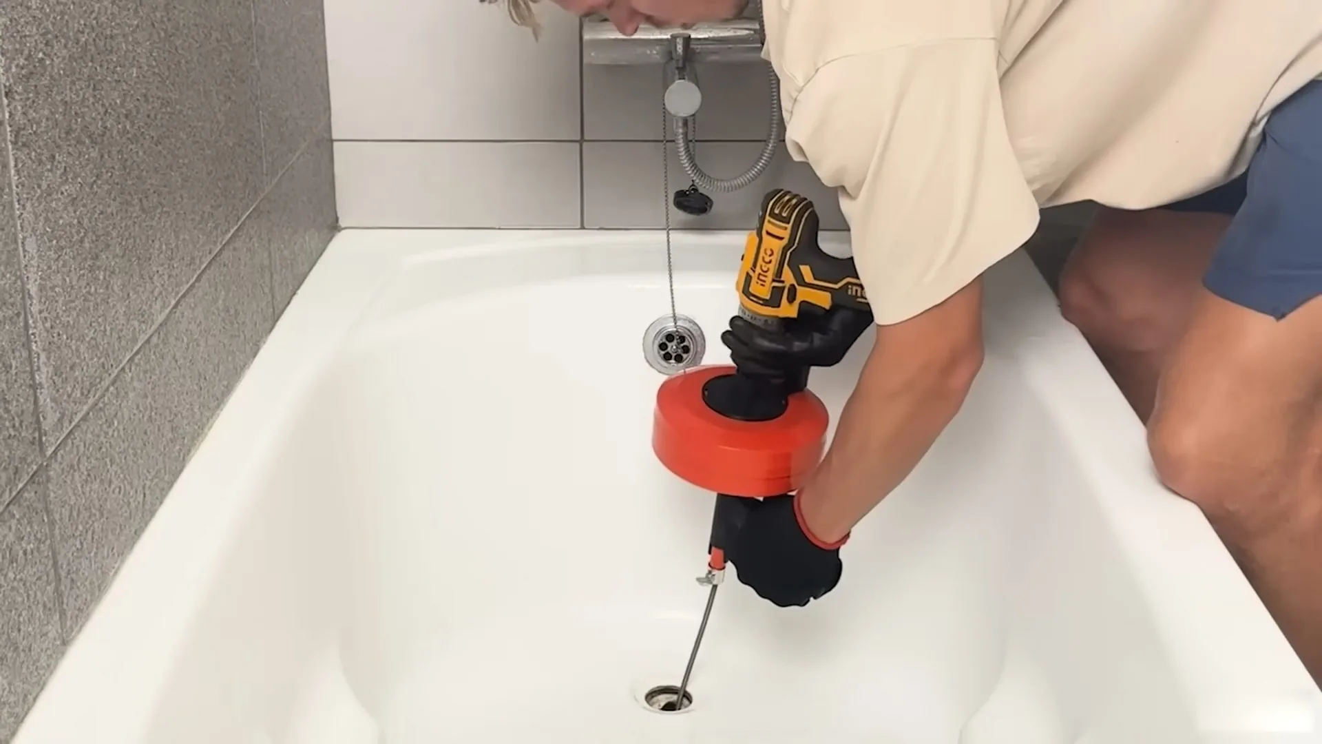 A person feeding a powered drain snake into the drain of a white bathtub.