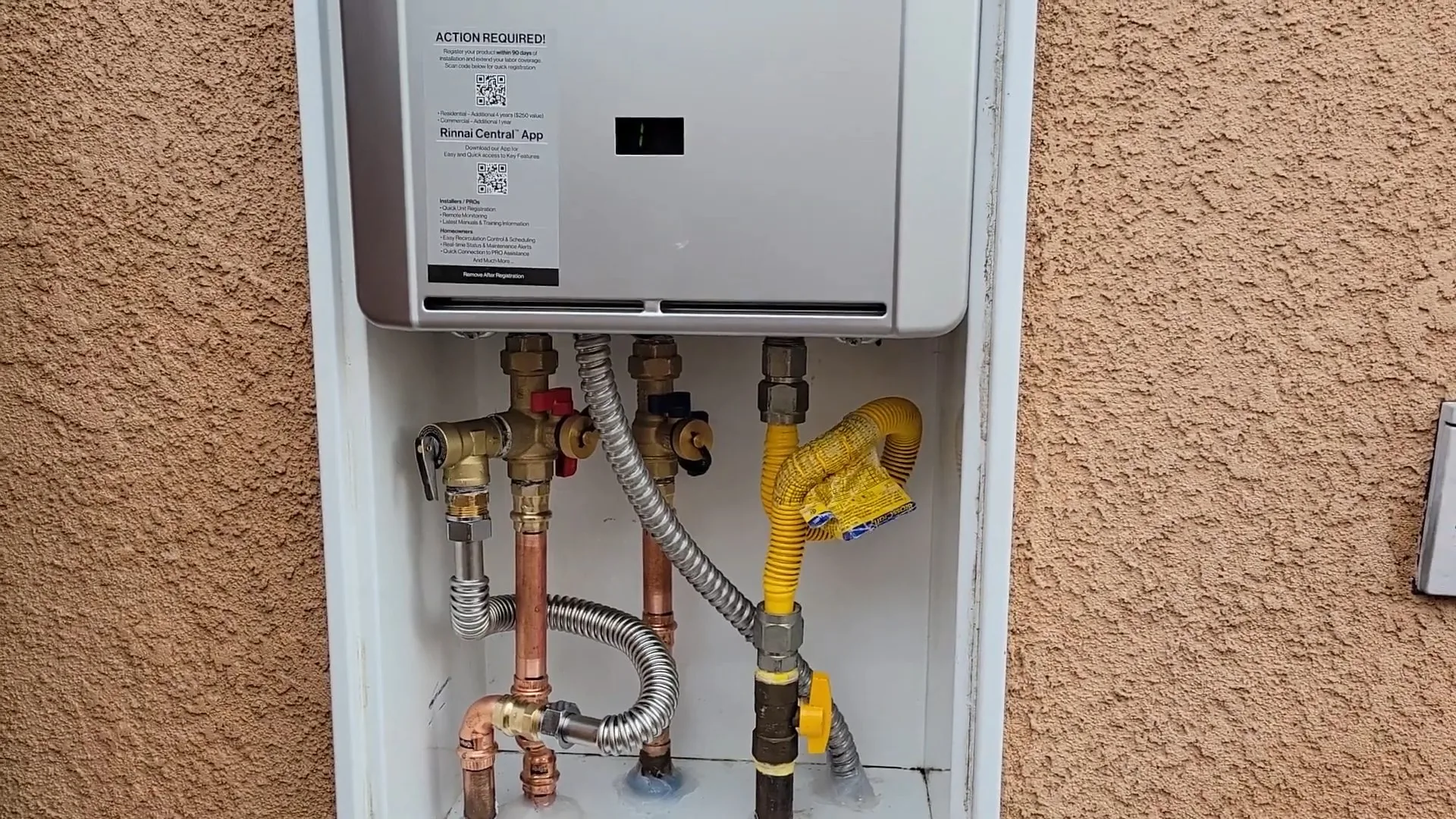 A wall-mounted tankless water heater in a recessed stucco cabinet with valves and a gas line below it.