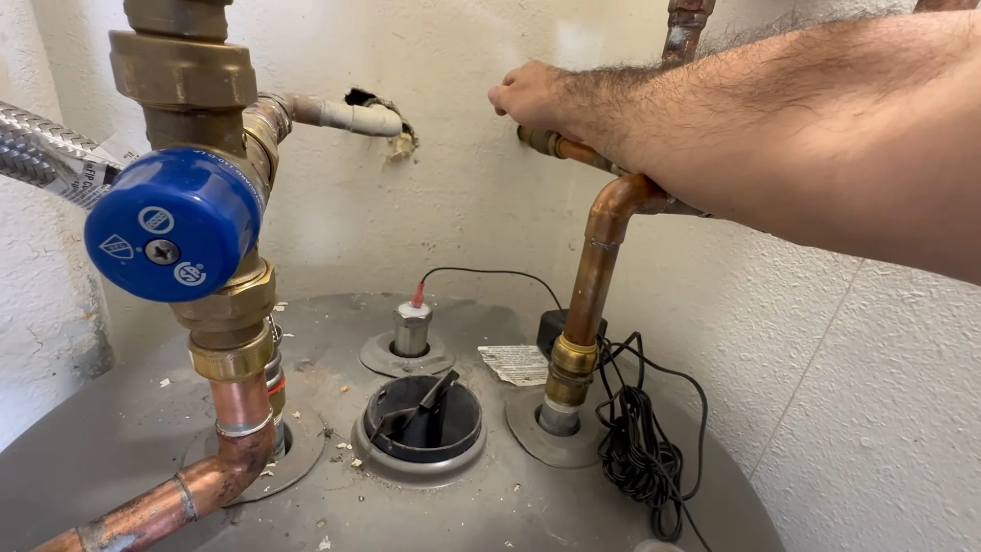 The top of a tank water heater with copper supply lines, a blue-handled valve and a hand reaching toward the outlet pipe.