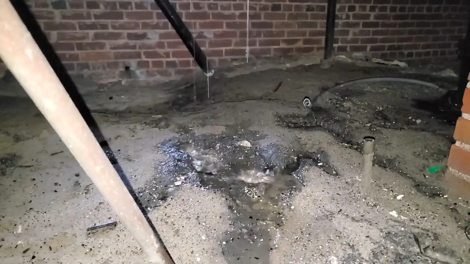 Wet debris and standing water on the floor of a crawlspace beneath a pipe, with a brick wall behind.