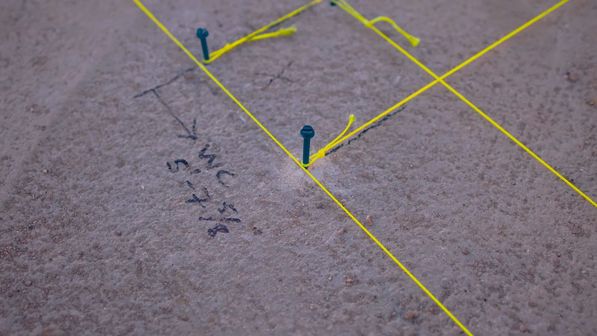 A floor surface marked with a yellow string grid, stakes and handwritten marker notes.